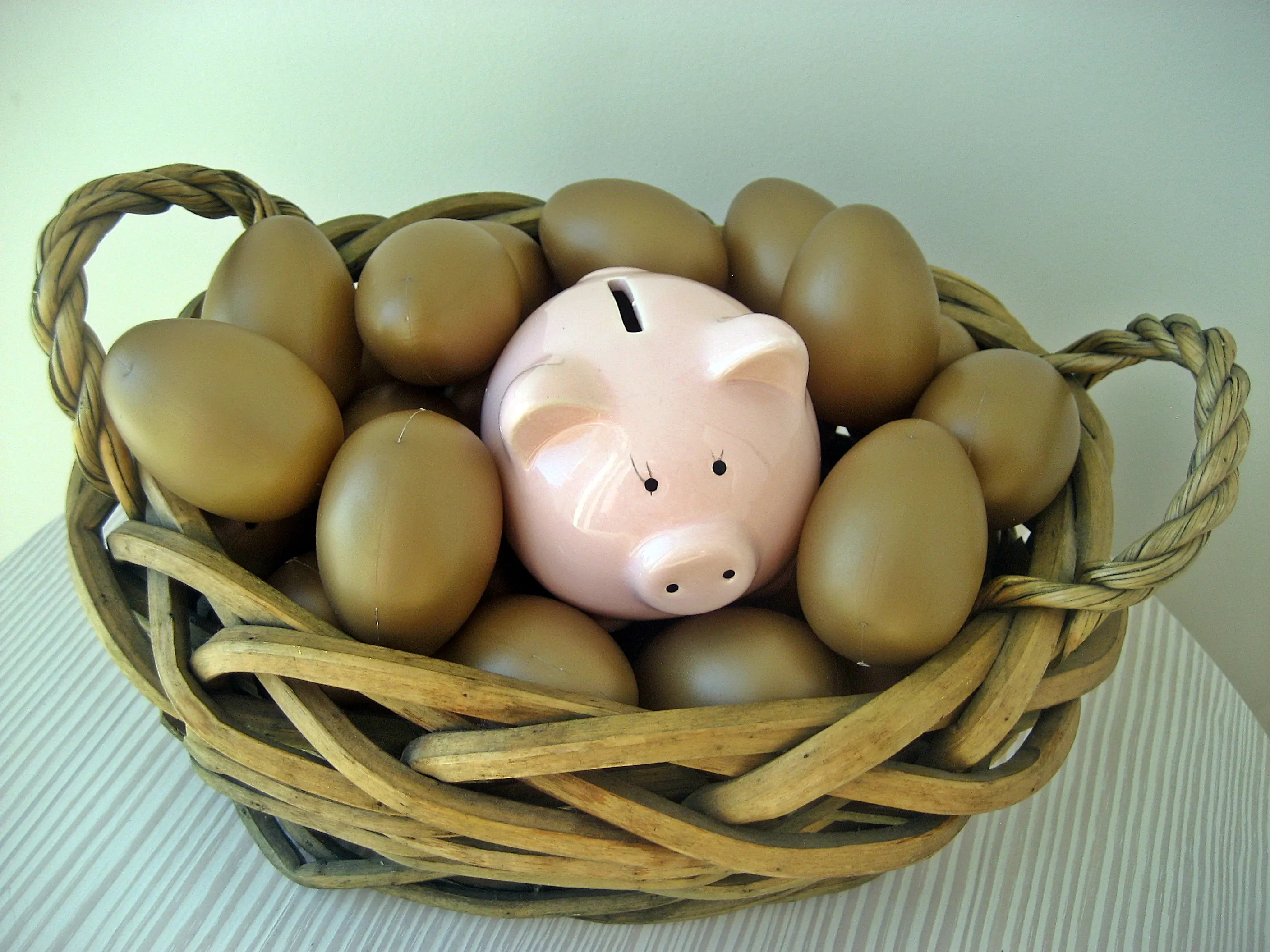 A woven straw basket with rope handles contains a pink ceramic piggy bank in the center, encircled by brown eggs